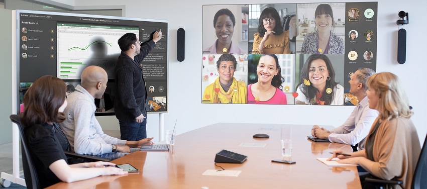 Two woman and three men are hosting a coordinated meeting in a large conference room and are joined by several virtual attendees on a projection screen in Front of Room view.