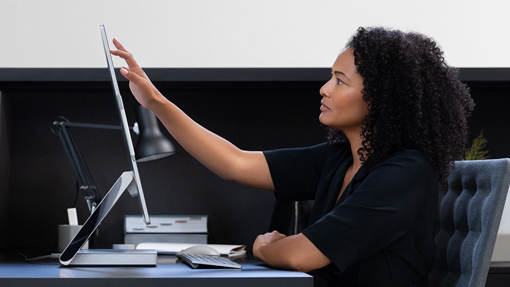 A woman at her desk reaches out to touch the Surface Studio 2 touchscreen display
