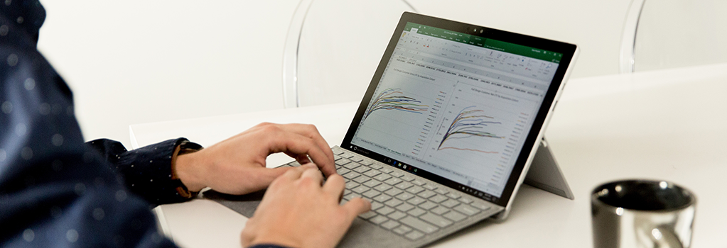 Close-up photograph of a person seated at a table working on a laptop. The laptop is displaying a graph in Excel.