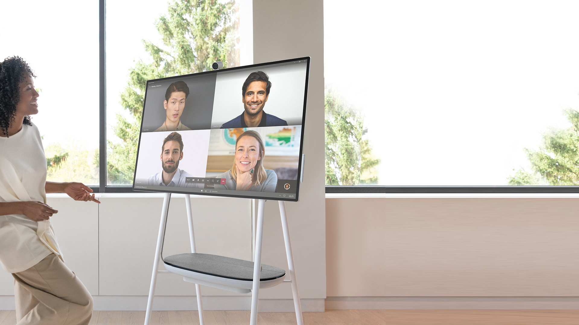 Eine Frau steht ungezwungen in einer Videokonferenz mit vier entfernt arbeitenden Teammitgliedern neben einem Surface Hub 2S