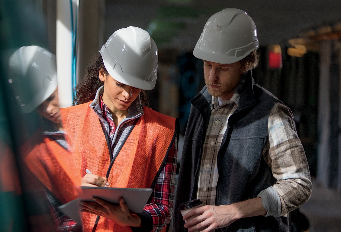 Photograph of two people wearing construction hard hats and gear talking inside a building that is under the final stages of construction. One of them is holding a tablet device.