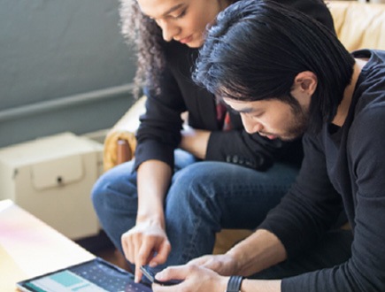 Photograph of two people sitting on a couch, one is using a tablet device, one is using a mobile device