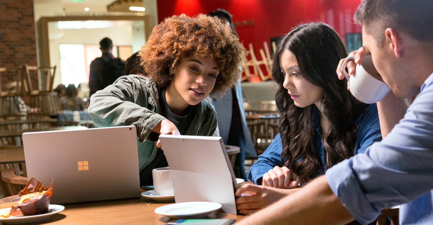 Three people seated at a table in a café with a Surface Book and a Surface Pro. One person is pointing at the screen of the Surface Pro while the others look on.