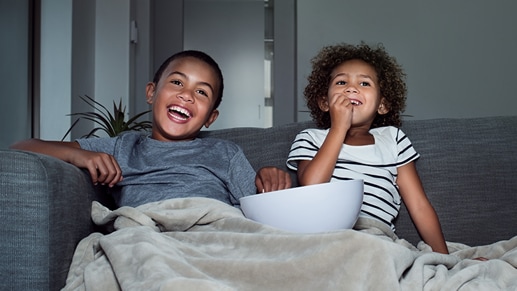 Two children share a bowl of snacks and a blanket while enjoying a movie at home.