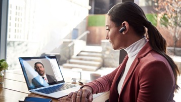 A person in business attire, wearing a wireless in-ear speaker, sits at a desk and appears to be participating in a video chat with someone who appears on her laptop screen.