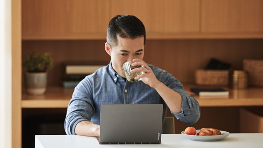 A person sits at a table sipping a drink while working on a laptop. A plate of food sits on the table next to the laptop.