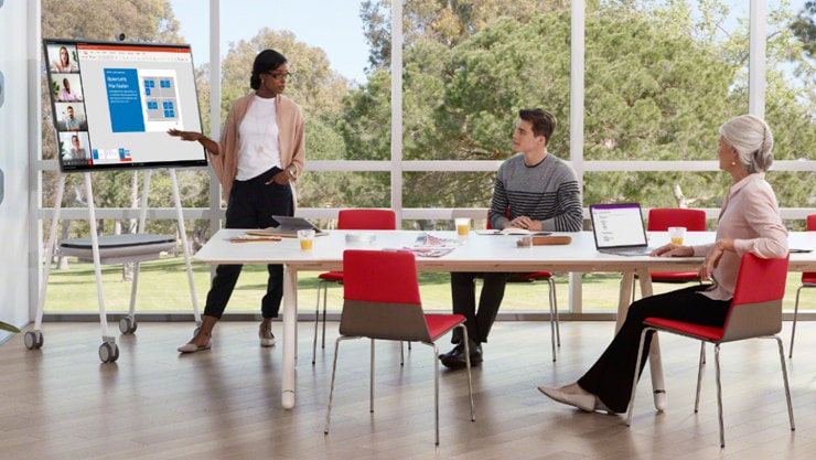 Team in einer Besprechung in einem Konferenzraum mit Fenstern. Eine Frau zeigt auf das Surface Hub 2S und präsentiert etwas