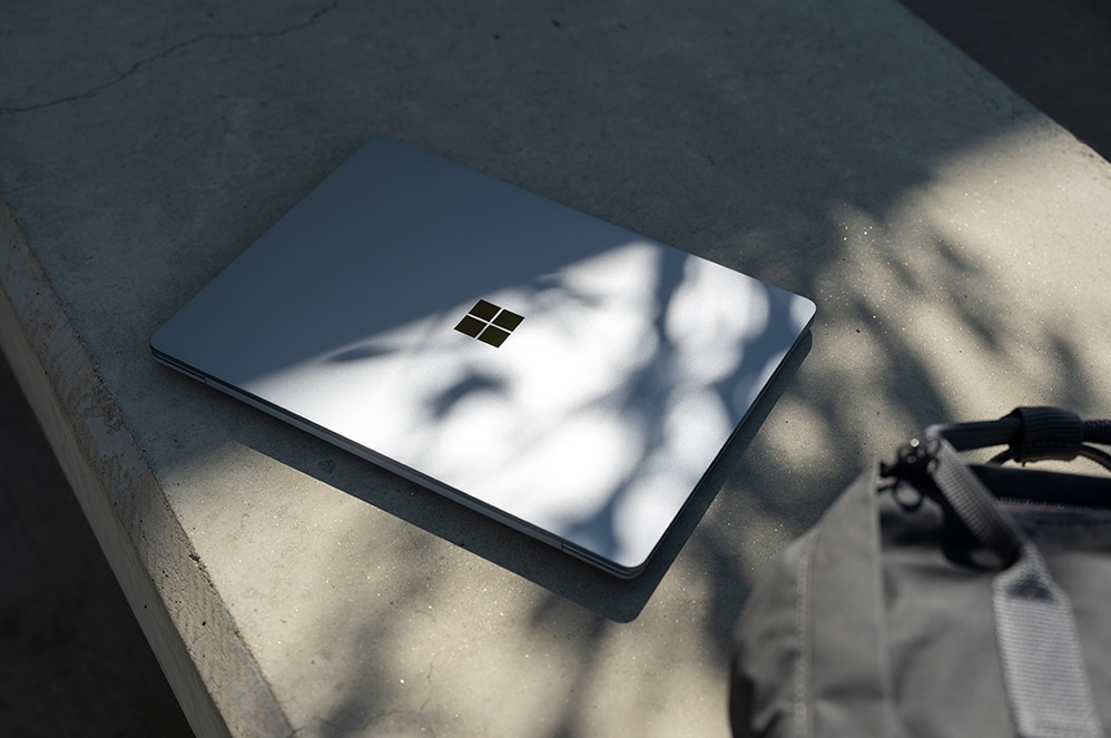 A Surface Laptop Go sits closed upon a concrete bench with a backpack nearby.