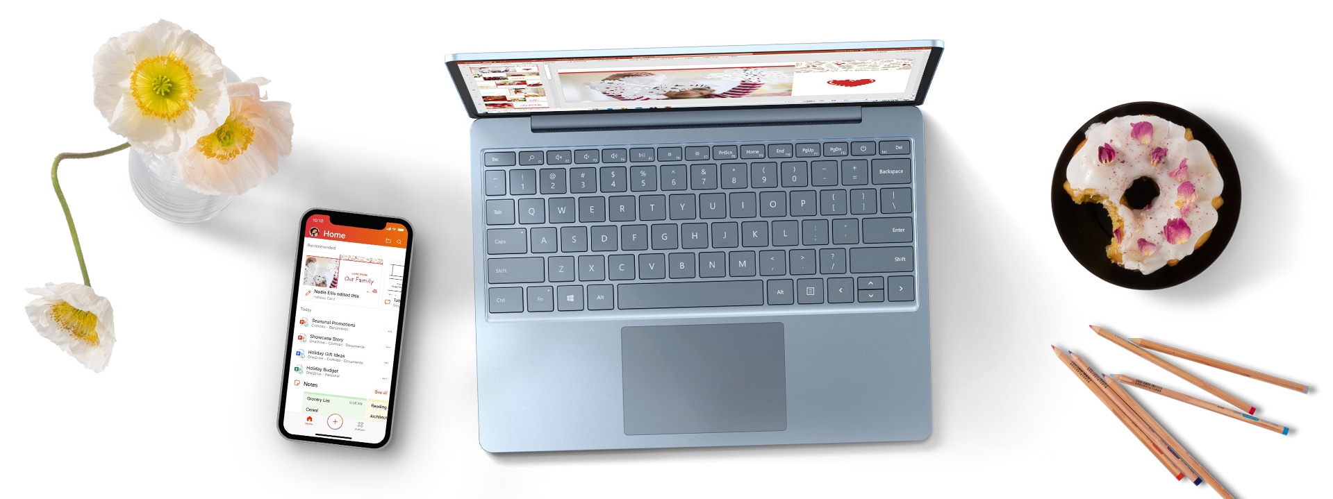 Surface Laptop Go auf einem Schreibtisch mit einem Handy, Blumen und einem Donut auf einem Teller