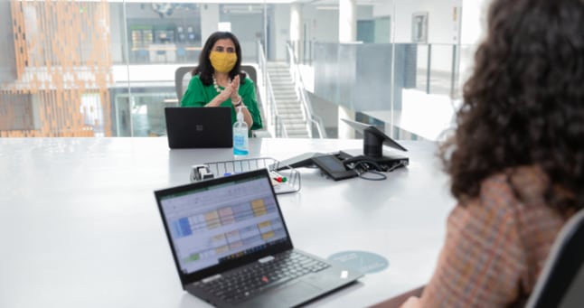 Two people in masks sitting at a desk with their laptops talking.