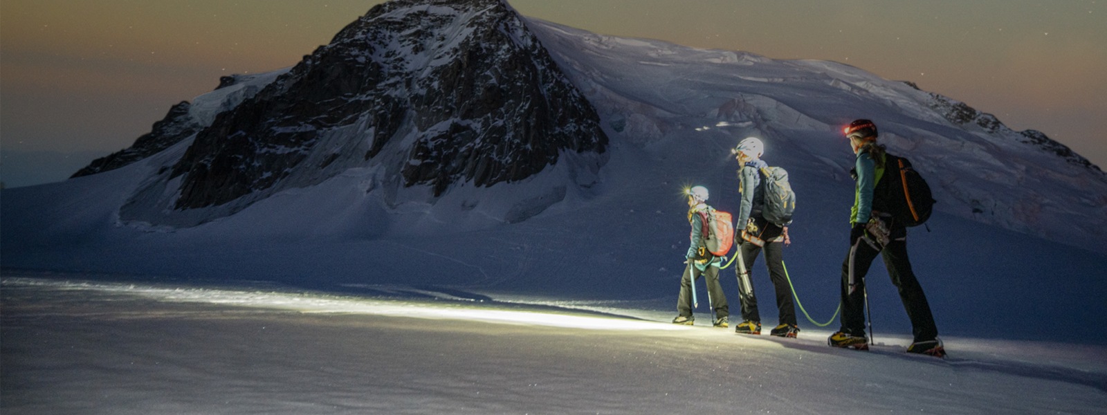 Drei Wanderer überqueren in den Bergen ein Schneefeld unter freiem Sternenhimmel.