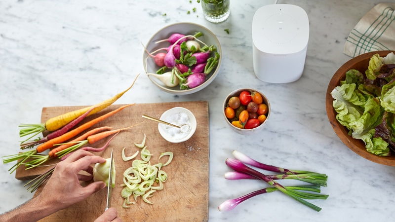 A person's hands chopping vegetables while the Sonos One in White is on the counter beside the cutting board
