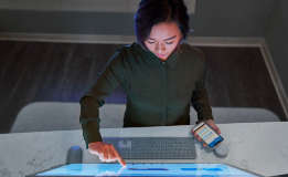 A person using a touchscreen desktop monitor while holding a mobile phone.