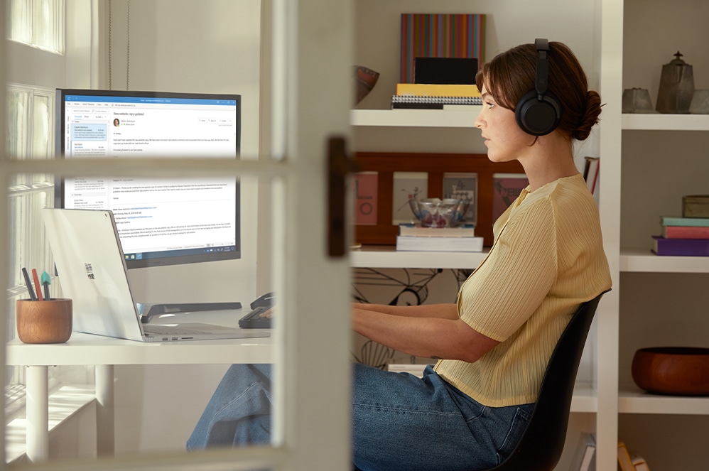 Person, die mit Surface Headphones+ am Schreibtisch sitzt und einen Computerbildschirm betrachtet.