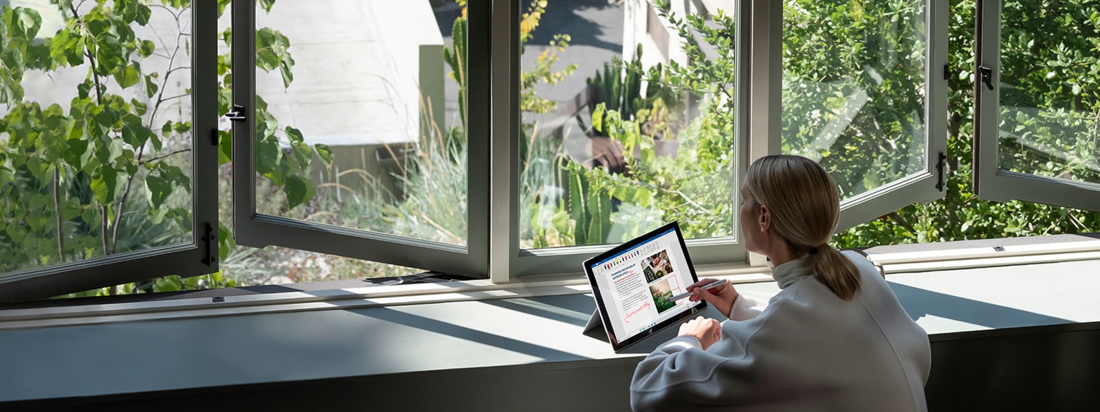A woman sitting near bright open windows with a view of green plants and working on a Microsoft Surface Laptop.