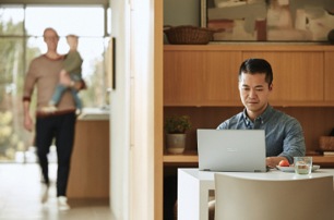 Person seated at a desk in a home office looking at a laptop, there is another person walking in the background and carrying a child