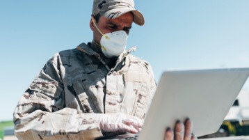 A member of the military in uniform and wearing a mask using a laptop.