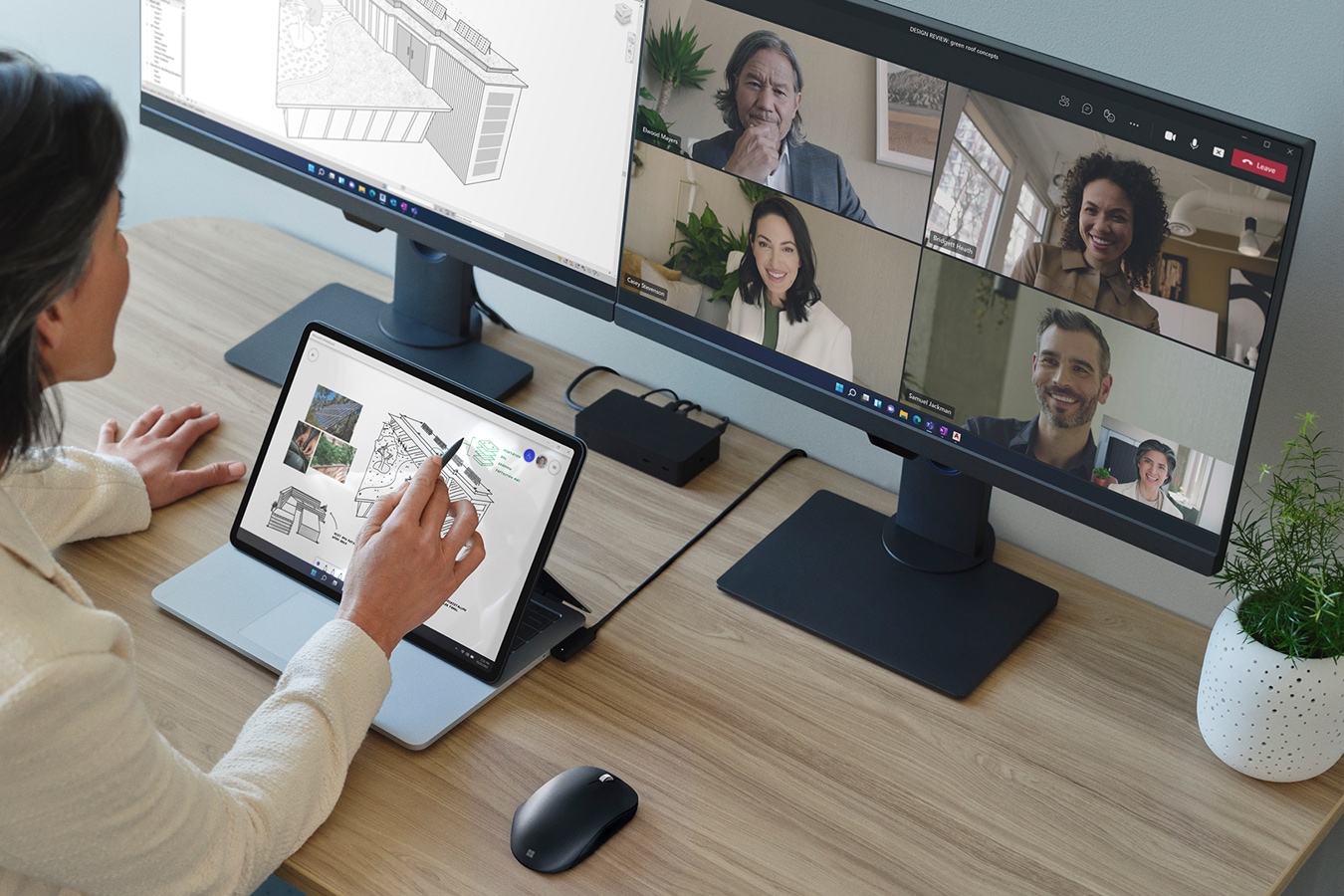 A person gesturing at the screen of Surface Laptop Studio for Business with a monitor in the background showing people in a Teams call.