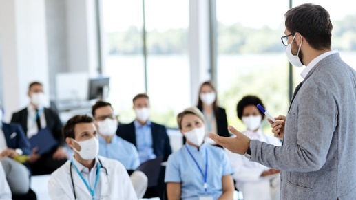 A person speaking in front of a group of people al wearing protective masks.