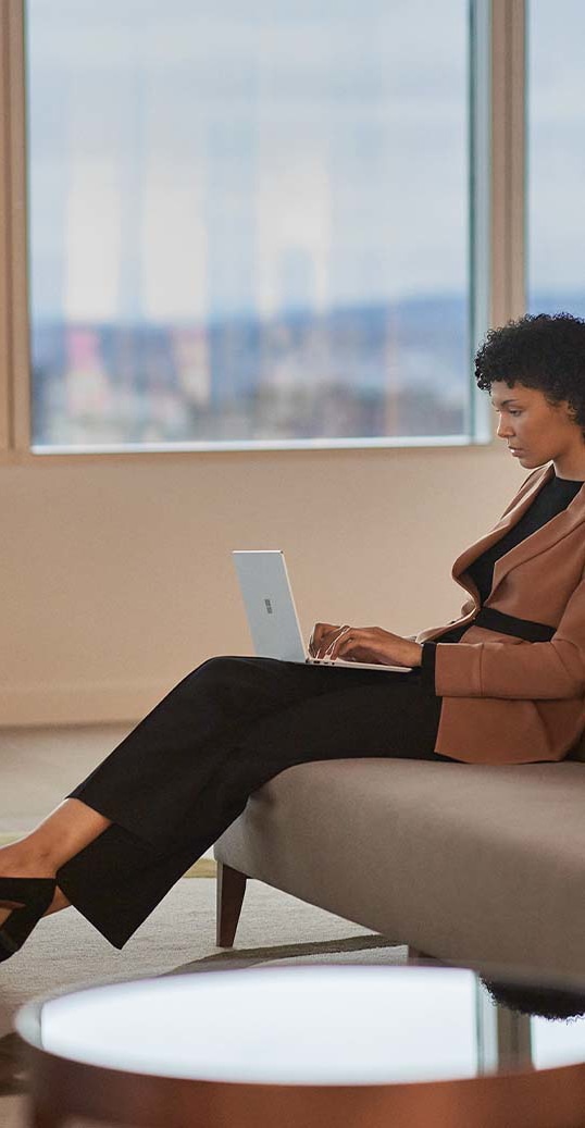 ผู้หญิงคนหนึ่งกำลังนั่งอยู่บนโซฟาในสำนักงานขณะที่พิมพ์บน Surface Laptop Go 2 ของตนเอง