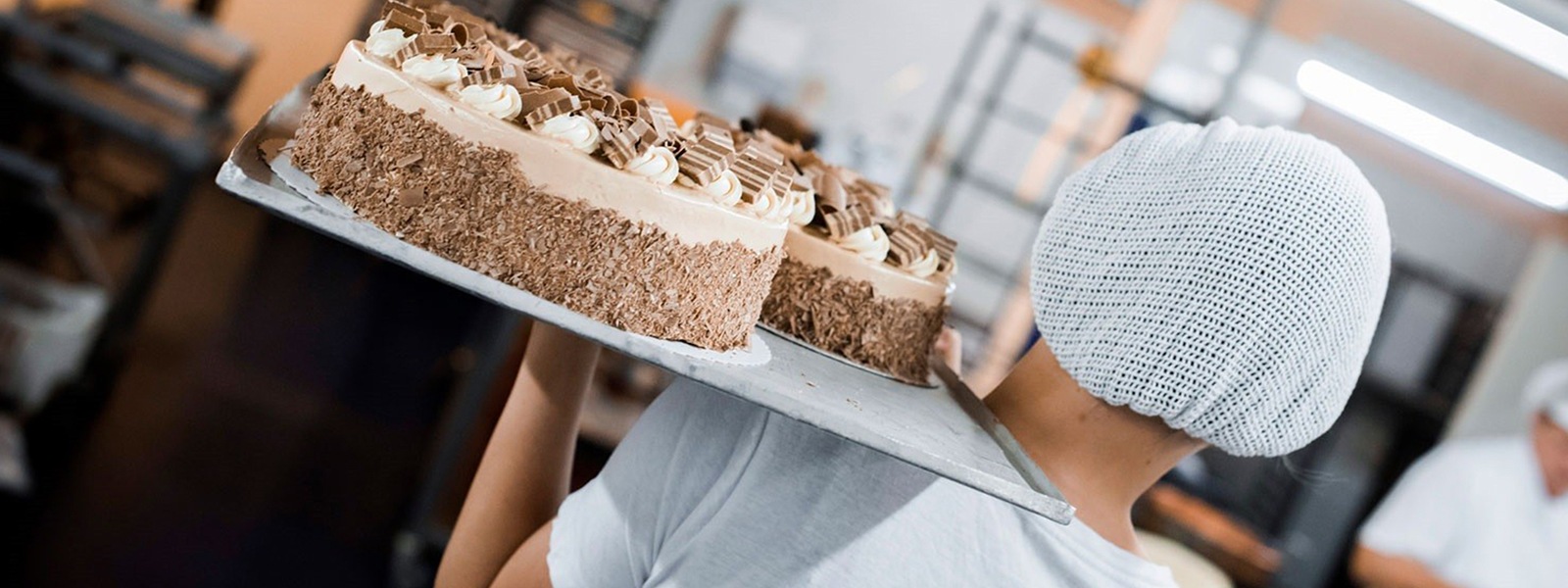 A worker carrying a large sheet pan with multiple cakes on their shoulder.