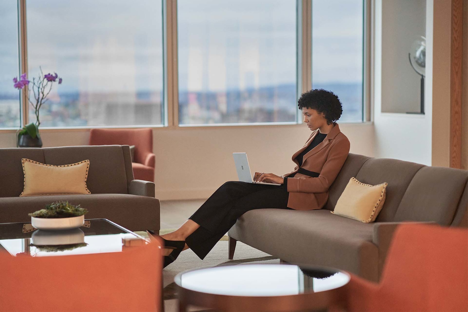 ผู้หญิงคนหนึ่งกำลังนั่งอยู่บนโซฟาในสำนักงานขณะที่พิมพ์บน Surface Laptop Go 2 ของตนเอง