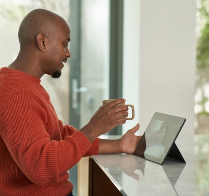 A smiling man in a red sweater sits in an open room holding a coffee mug and watching a tablet screen.