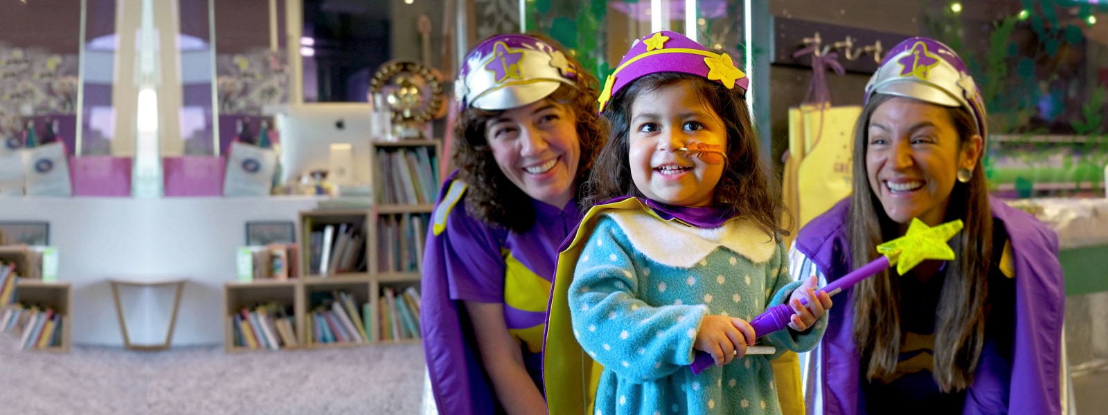 Two adult volunteers in costumes laugh alongside a small child dressed in a costume holding a wand