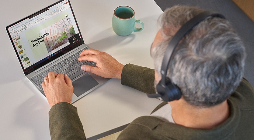 戴著 Microsoft 時尚無線耳機麥克風的男人坐在桌前，使用 Surface Laptop Go 2 出席 Microsoft Teams 會議的俯視圖