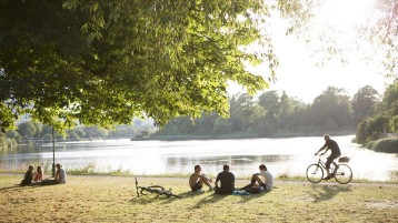 A park with a lake, people relaxing in the grass and someone biking on a trail.