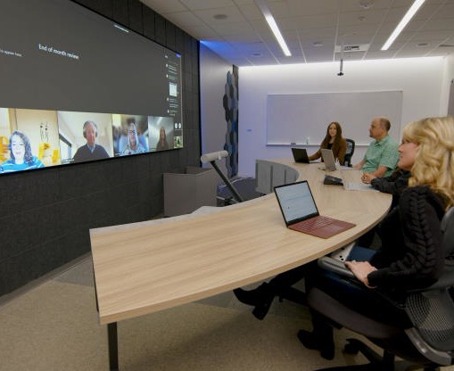 A Microsoft team meets at a curved desk facing a screen on the wall, allowing them to see everyone in the meeting equally.