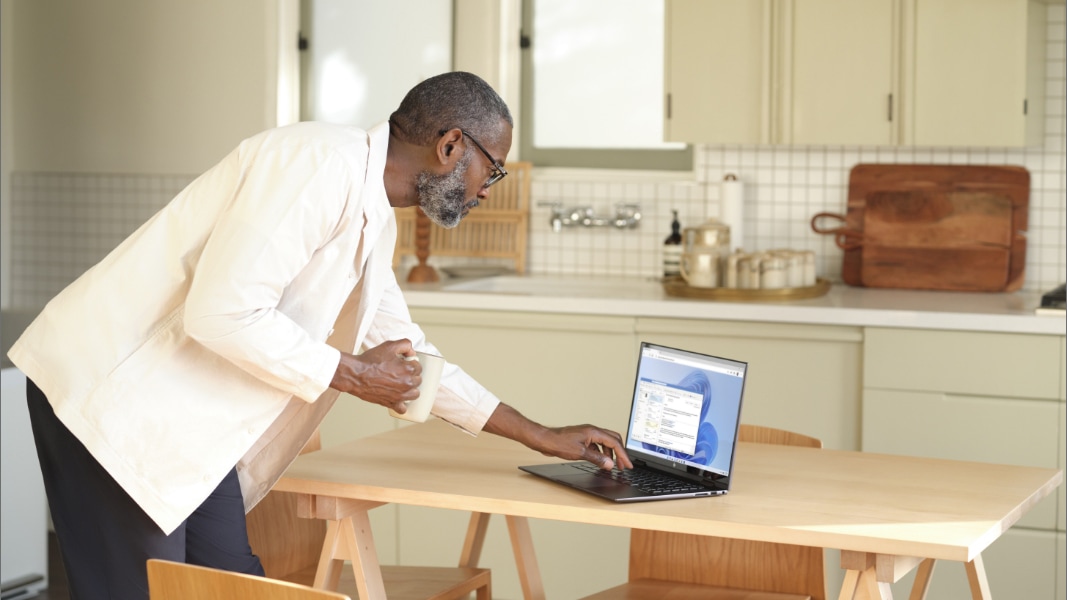 A person leaning over a kitchen table to type on a laptop that is running Outlook.