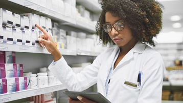Gesundheitsexpertin in einer Apotheke, die auf ein Medikament in einem Regal zeigt und Informationen auf dem Tablet abliest