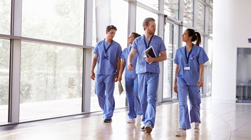 Vier Mitarbeiter im Gesundheitswesen in Arbeitskleidung, die in einer Gesundheitseinrichtung zu Fuß gehen. Einer von ihnen trägt ein Tablet