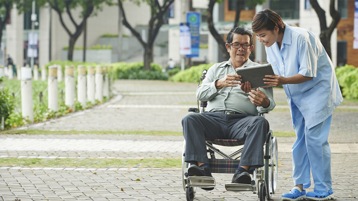 Ein Mann im Rollstuhl und eine Frau, die neben ihm steht, schauen gemeinsam auf ein Tablet