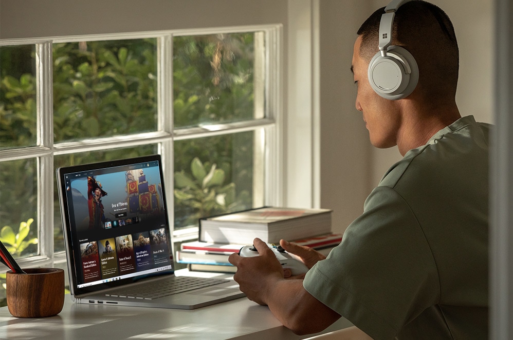 رجل يمارس إحدى ألعاب Xbox على جهاز Surface Book 3 باستخدام وحدة تحكم Xbox أثناء ارتداء سماعات الرأس Surface