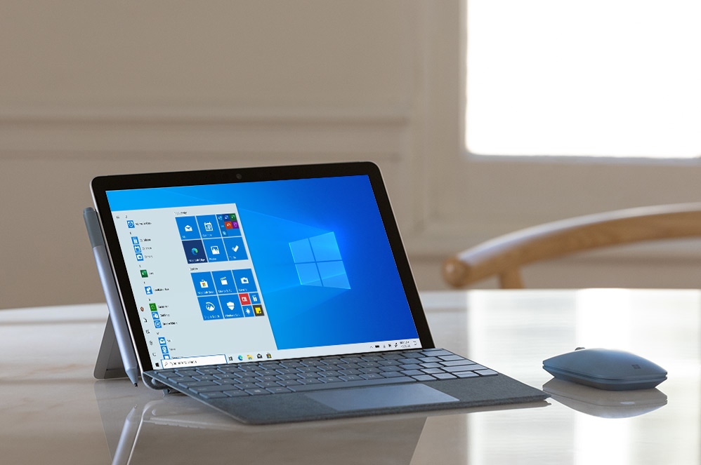 Komputer Surface Go 2 z wyświetlonym ekranem startowym Windows 10 Gmunk znajdujący się na biurku obok myszy Mobile Mouse