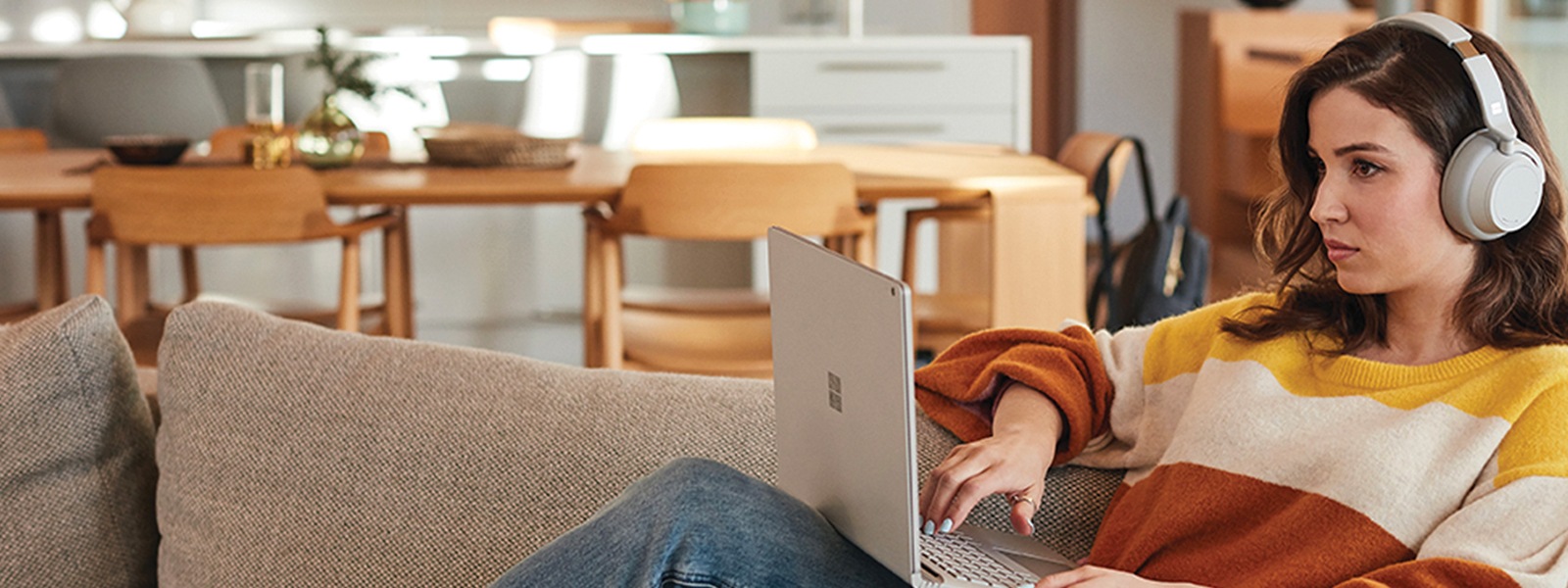 一名女子在家躺在沙發上使用 Surface Laptop 和 Surface Headphones。