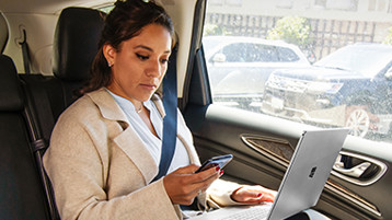 A person sitting in the back seat of a car using a laptop and looking at a mobile phone