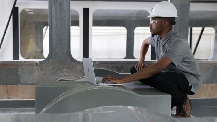 Person wearing a hard hat crouches down to use a laptop in a construction environment.