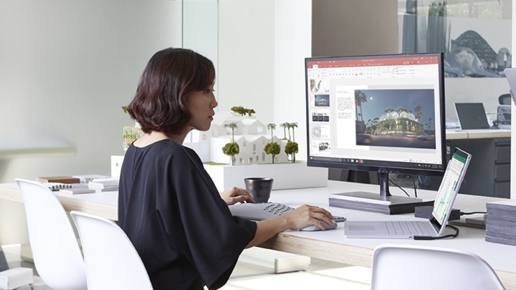Person sits alone at a desk working on a PowerPoint presentation on a large desktop monitor.