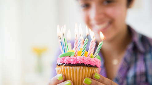Photo credit: JGI/Jamie Grill/Getty Images. A smiling woman holding a cupcake with colorful birthday candles
