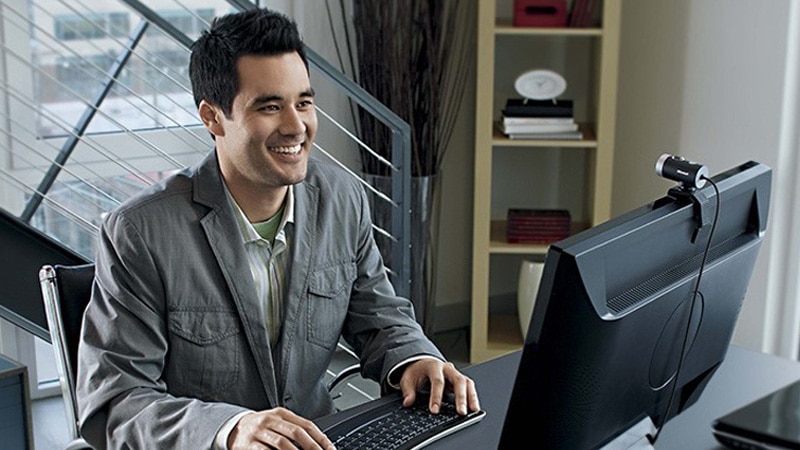 Un hombre sentado en un escritorio con un teclado, frente a un monitor de computadora con una cámara web de cine lifecam montada en él