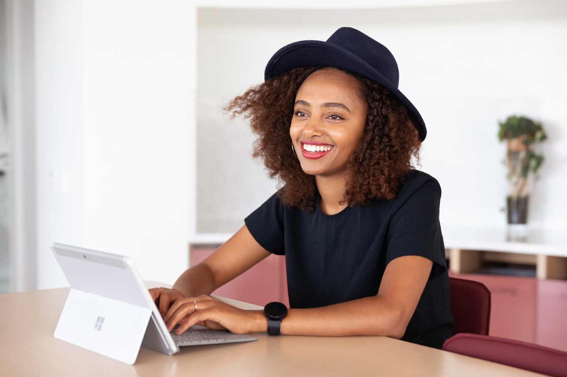 A person wearing a hat smiling and typing on a Surface device.
