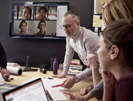 A group of people having a meeting in a conference room looking at architectural blueprints while other team members are dialed in through a Teams video call being displayed on a screen on the wall behind them.