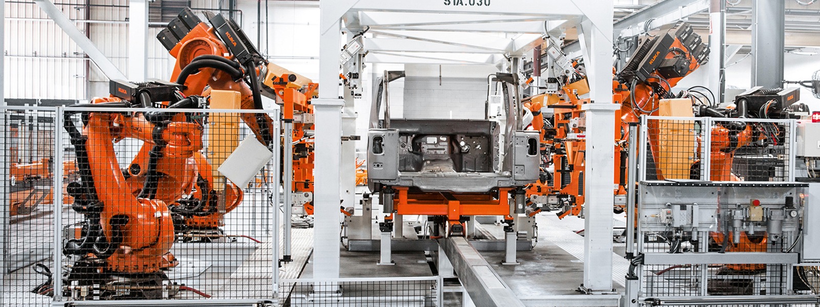 Das Bild zeigt eine Produktions-Straße, wobei auf beiden Seite orangene KUKA Roboter arbeiten.