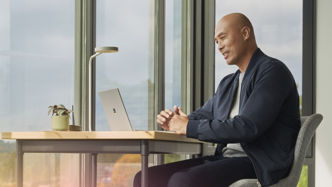 A person sitting at a desk with their hands folded participating in a video call on a laptop.