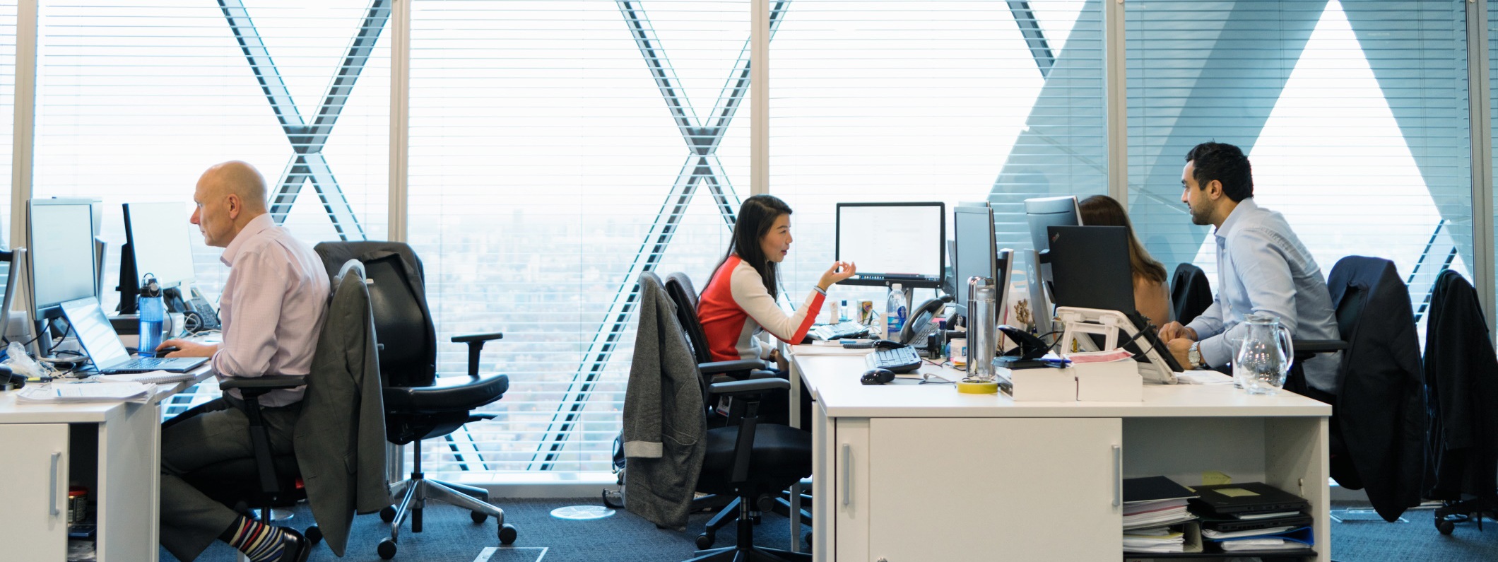 A female and male office worker in conversation, facing each other at shared workstation in open office setting. 