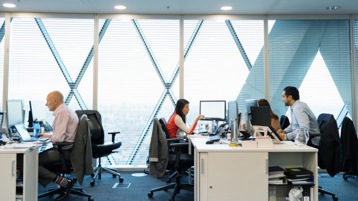A female and male office worker in conversation, facing each other at shared work station in open office setting. 