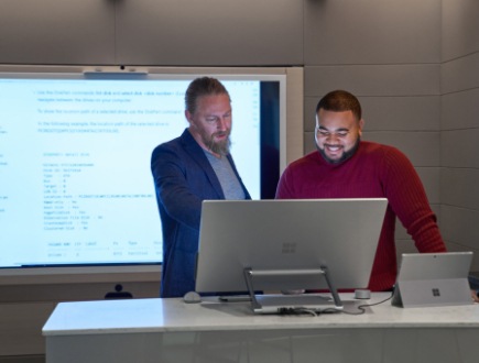 Two people working together at a desktop computer in front of a large screen displaying code.